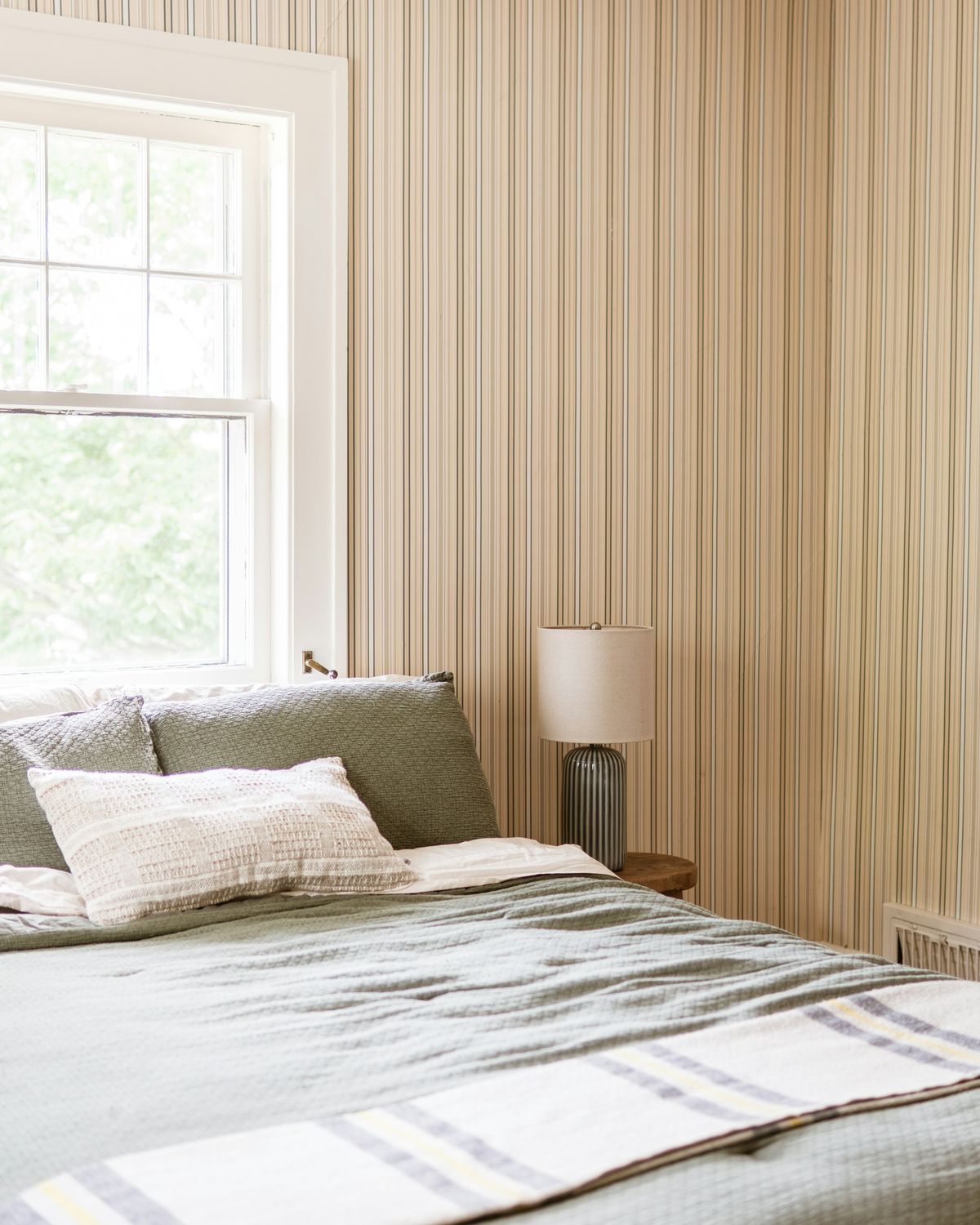 Bedroom with fine vertical striped wallpaper, sage-green bedding and a window letting in soft daylight