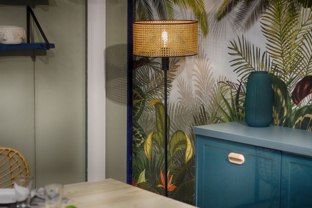 Dining corner with a tropical leaf wallpaper mural, a rattan floor lamp and a teal cabinet