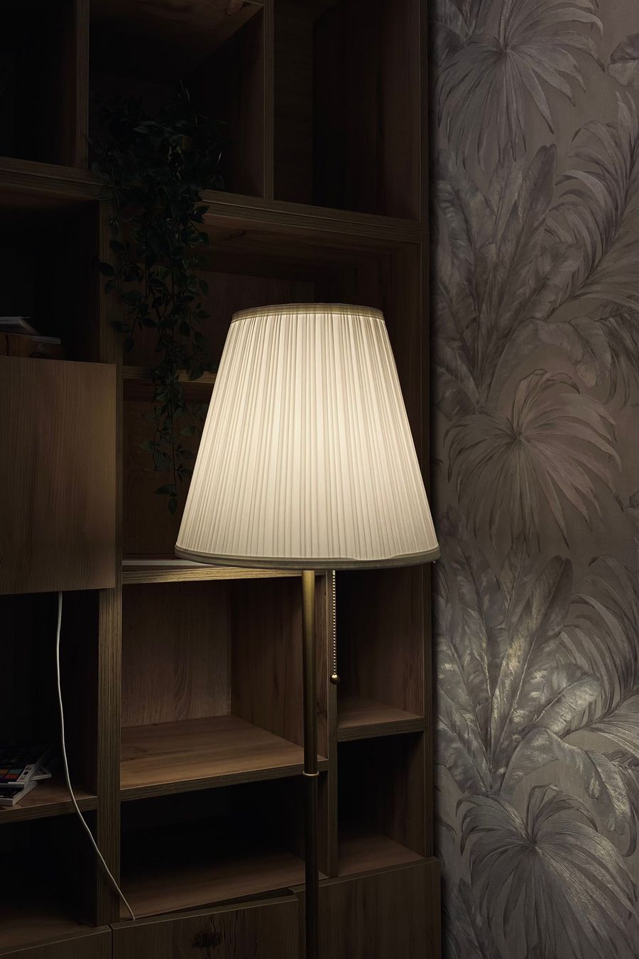Pleated floor lamp glowing beside a tonal palm-leaf wallpaper next to wooden shelving