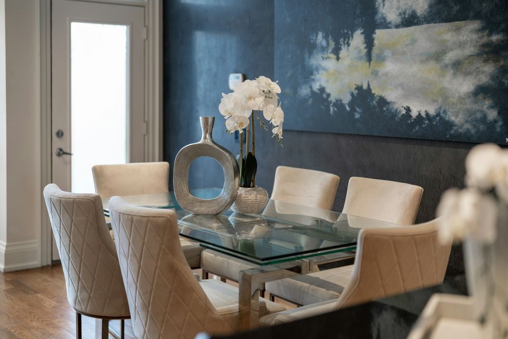 Dining room with a large blue sky-and-cloud wall mural behind a glass table and cream upholstered chairs