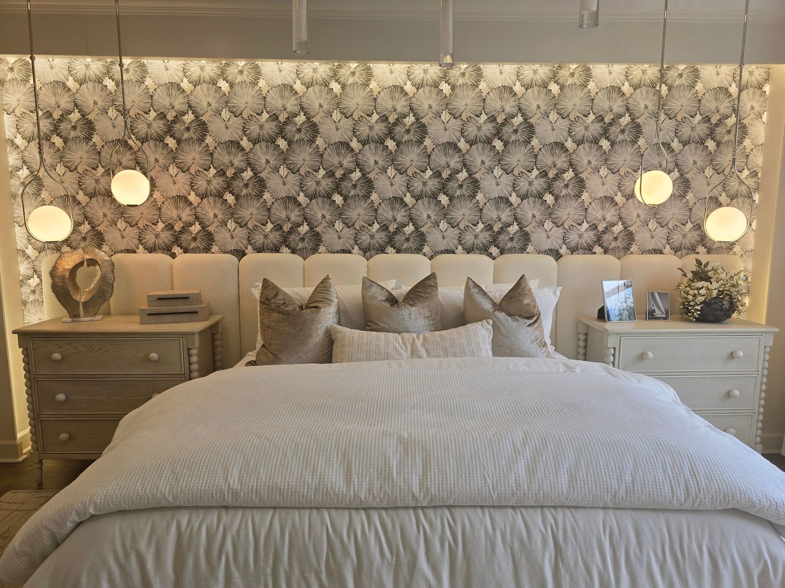 Bedroom feature wall in a textured botanical wallpaper behind an upholstered headboard, lit by four globe pendant lights
